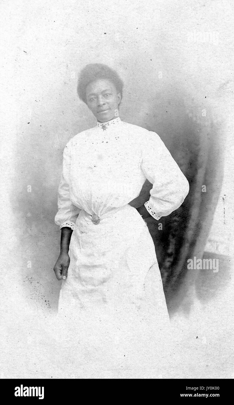 Portrait of an African American woman standing in front of a curtain, her left arm is folded behind her back and her right arm is by her side, she is wearing a light colored long dress that is cinched at the waist with a pendant, Atlantic City Post Card Souvenir, Myers - Cope Co, 1635, 1918. Stock Photo