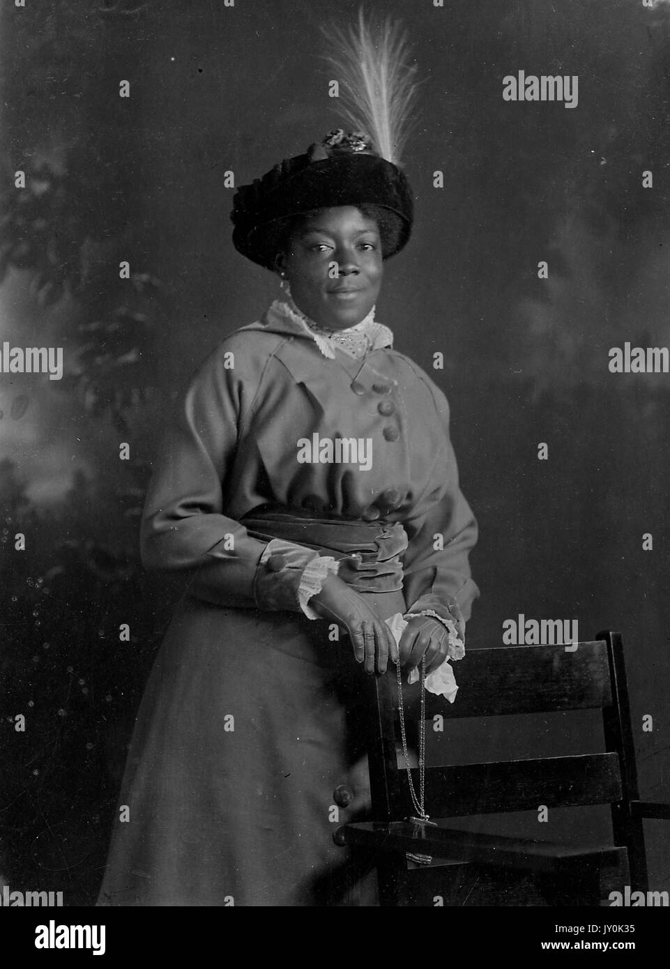 Portrait of an African American woman named Mattie E Bradford, she is wearing a long dress that is cinched at the waist, she is wearing a light colored shirt underneath, she is wearing gloves and a dark colored hat with a feather, her hands are resting on the back of a chair that is placed in front of her, a necklace dangles in her hands, she is smiling, The Electric Studio, 162 Thames St, Newport RI and Other Cities, 1915. Stock Photo