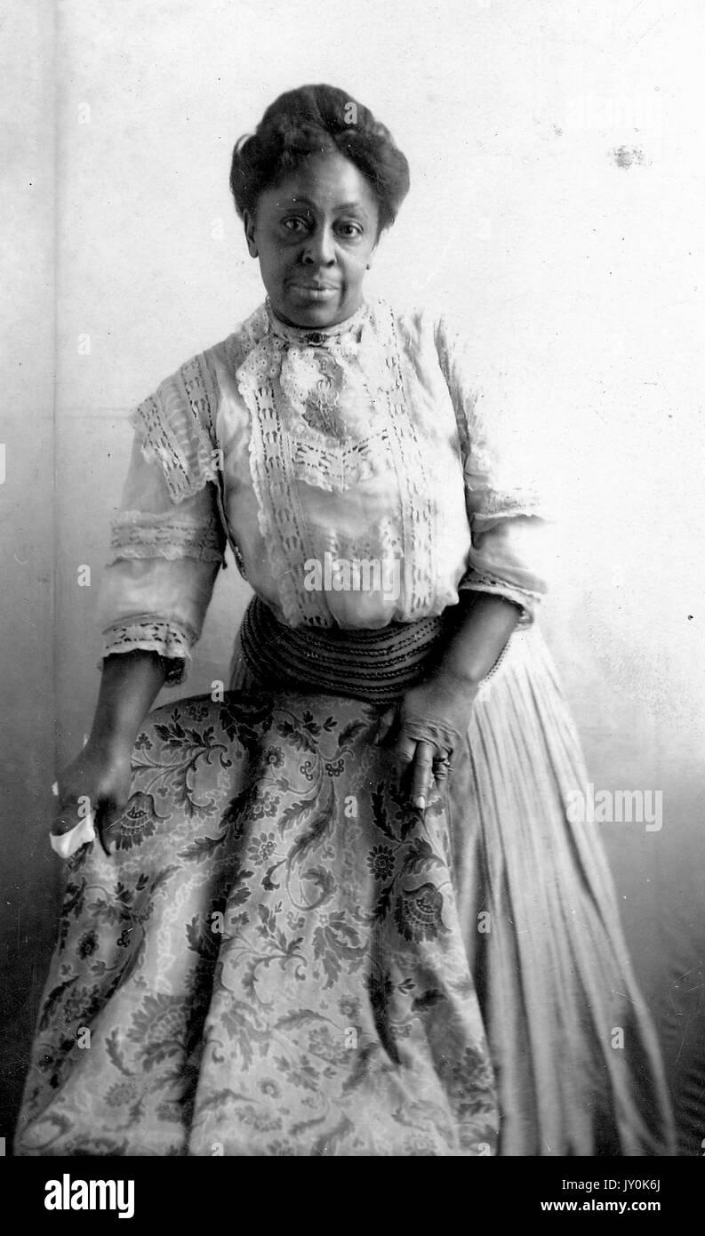 Three quarter length standing portrait of mature African American woman, wearing light lace blouse with dark skirt, standing next to and grasping chair, neutral expression, 1915. Stock Photo