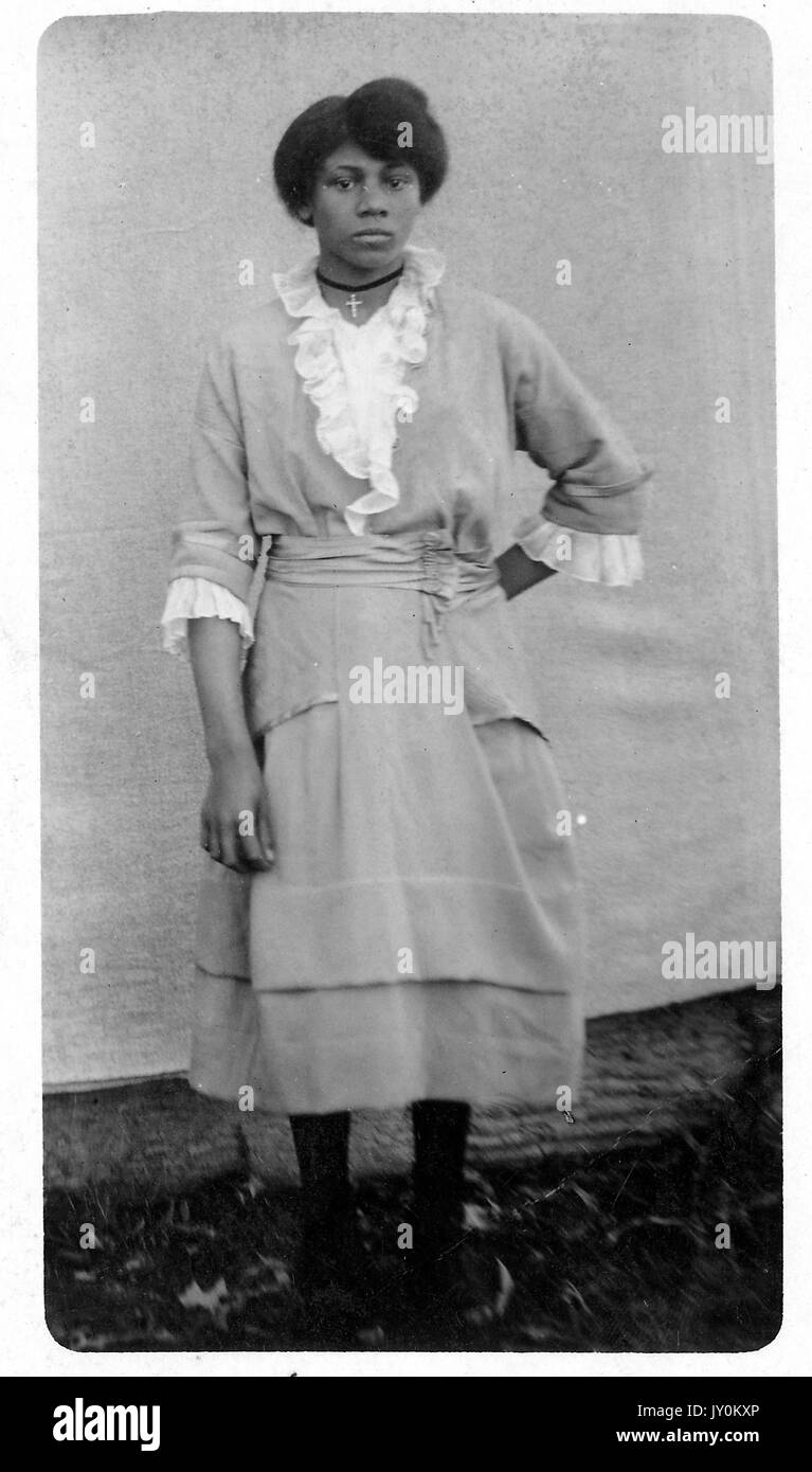 Full length standing portrait of young African American woman, wearing light dress with lace detail and necklace with a cross, standing outdoors next to a wall, neutral expression, 1915. Stock Photo