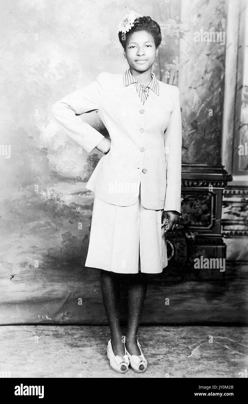 Full-length of African American woman, wearing a white dress and a white jacket, standing with one hand on her hip, with a flower in her hair, standing in front of a backdrop, with a serious facial expression, 1920. Stock Photo