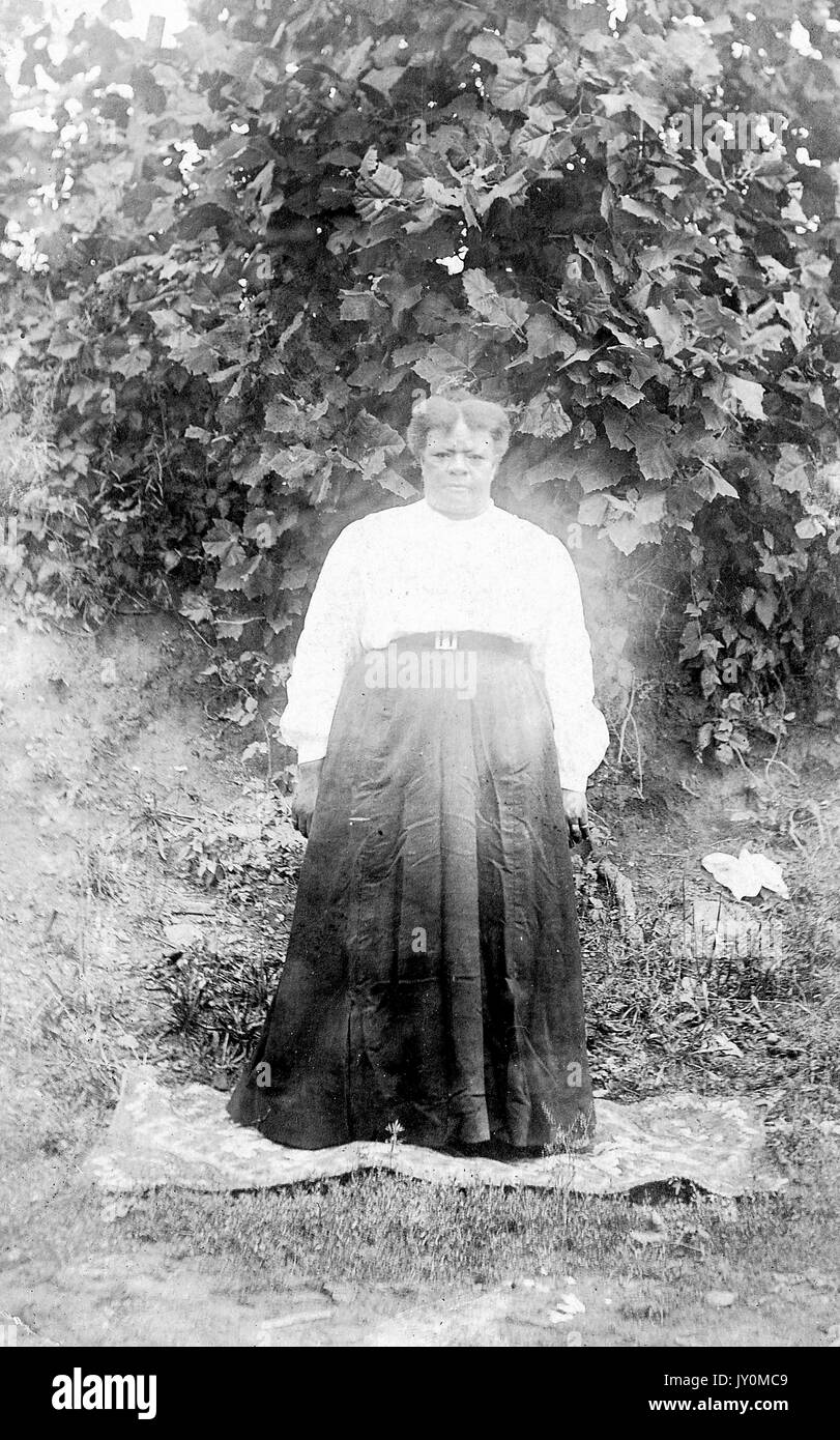 Full-length portrait of African American woman, standing on a blanket in front of a large bush in the grass, wearing a dark dress, arms by her side, with an angry facial expression, 1920. Stock Photo
