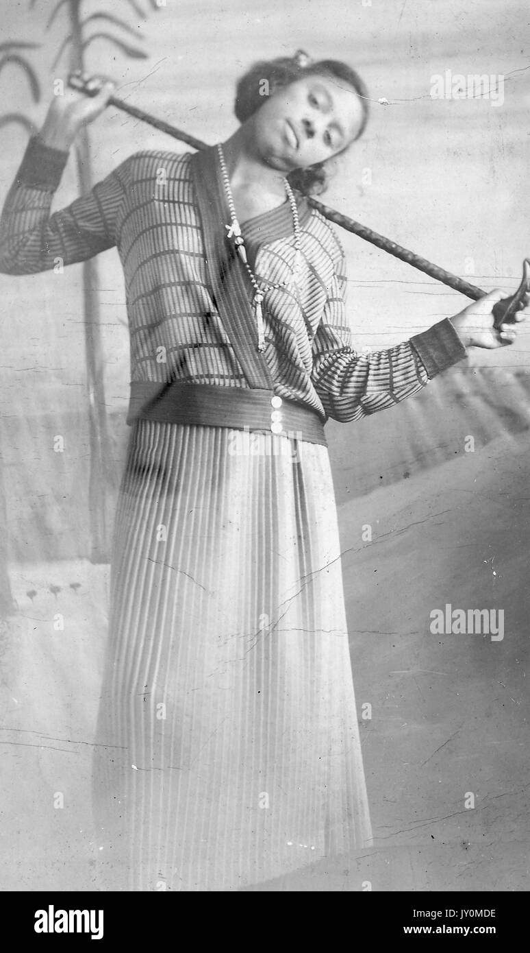 Three-quarter length portrait of African American Woman, standing in front of a backdrop, wearing a striped patterned dress, holding a dark wooden cane behind her neck with both hands, leaning her her head to her left, with a serious facial expression, 1920. Stock Photo