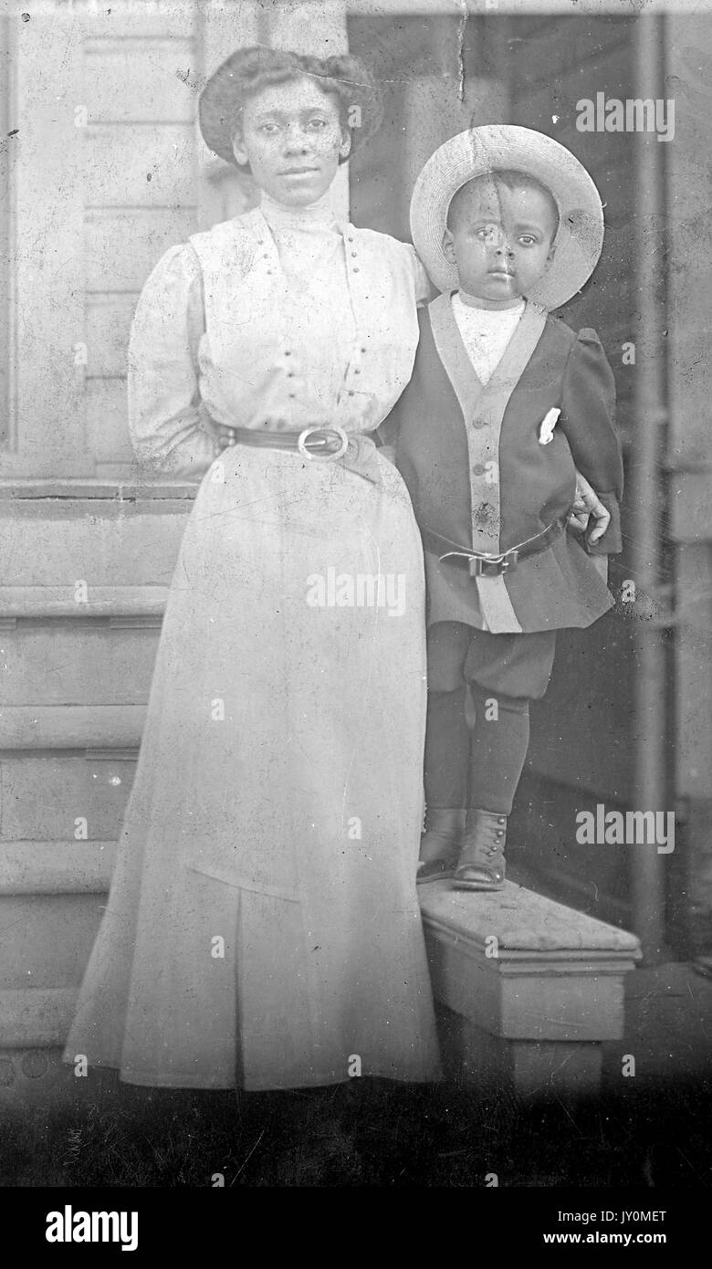 Full-length portrait of African American woman and boy, both standing, the woman's arm around the boy, both with serious facial expressions, the woman wearing a white dress and a dark belt around her waist, the boy wearing a dark cardigan and a hat, the boy standing on a ledge to meet the woman's height, 1920. Stock Photo