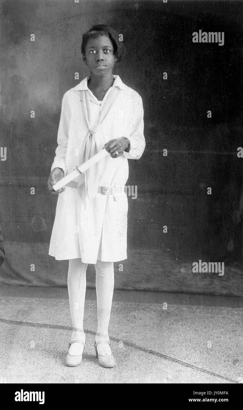 Full-length portrait of a young African American woman, wearing a white dress and white stockings, standing in front of a backdrop, holding a diploma rolled up in her hands, with a serious facial expression, 1929. Stock Photo
