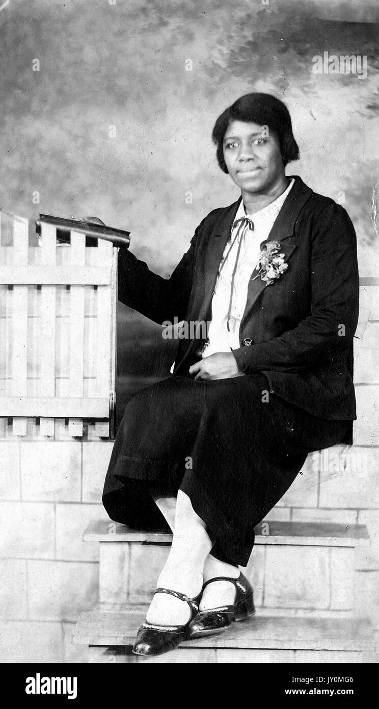 Full-body portrait of African American woman, sitting on steps in front of a backdrop, wearing a dark jacket and a dark dress, with a floral corsage on her lapel, resting a book on a fence with her right hand, resting her other hand in her lap, with a serious facial expression, 1920. Stock Photo