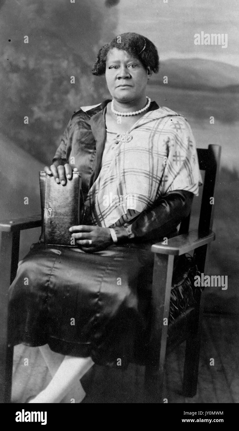 Full-body portrait of an African American woman, seated in a wooden chair, standing a purse on her lap, wearing a white and dark patterned dress, wearing a pearl necklace, with an angry facial expression, 1920. Stock Photo