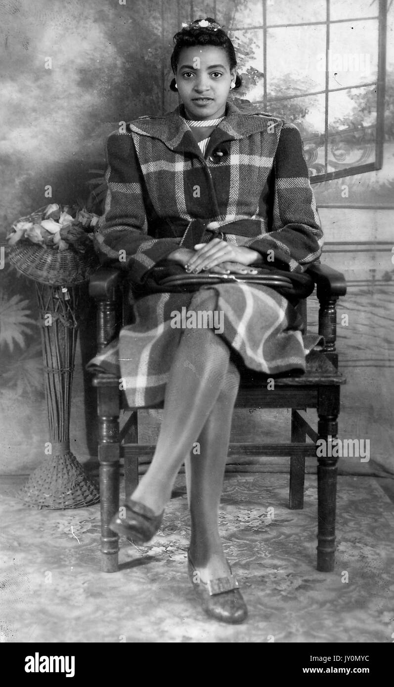 Full-body portrait of an African American woman sitting in a wooden chair, with her hands folded on her lap, legs crossed, wearing a large dark coat, seated in front of a backdrop, with a blank facial expression, 1920. Stock Photo