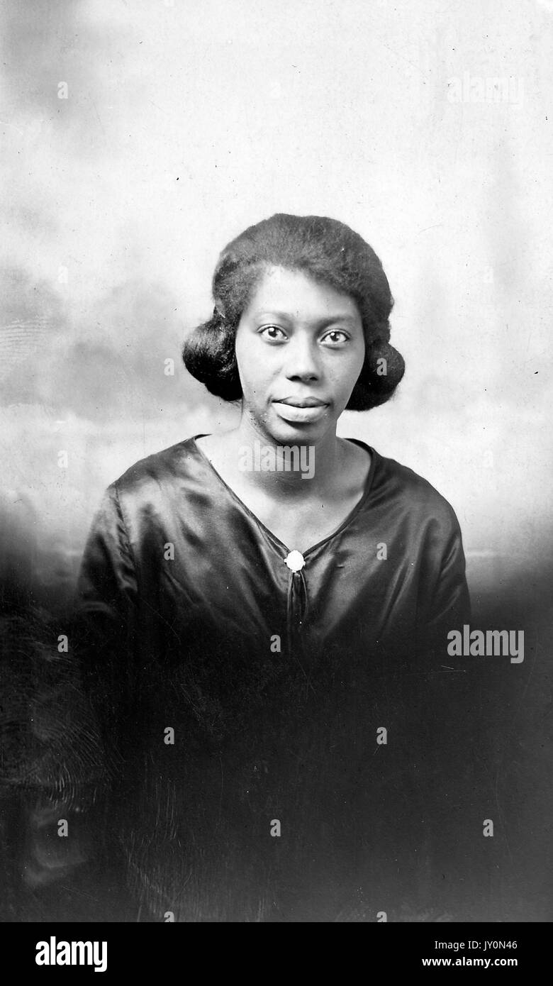 Half-length portrait of an African American woman, wearing a dark blouse, in front of a backdrop, with a serious facial expression, both arms by her side, as she stands, 1920. Stock Photo