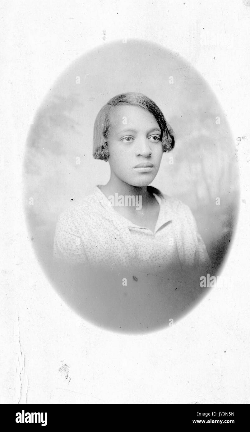 Oval head shot portrait of an African American woman, wearing a white blouse, with a serious facial expression, in front of a backdrop, 1920. Stock Photo