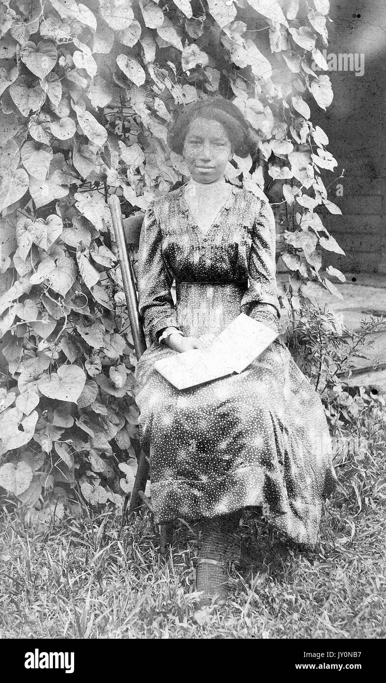 Full body portrait of an African American woman seated with an open book in on her lap, wearing a dark dress, with a closed-mouth smile facial expression, sitting on a wooden chair in the grass in front of a hedge, 1920. Stock Photo