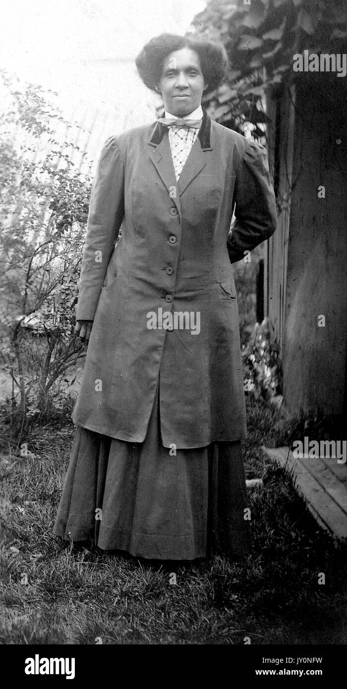 Full body portrait of an African American woman standing on grass, with one arm by her side and the other behind her back, wearing a dark dress and a dark coat over it, wearing a bowtie, with a serious facial expression, 1920. Stock Photo