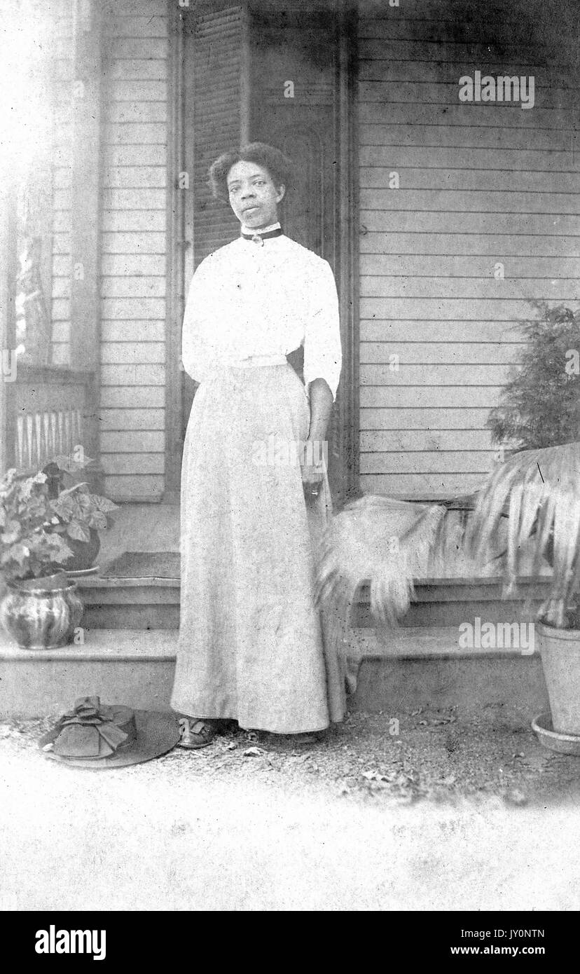 Full-body portrait of African American woman standing in front of a porch, wearing a white dress, with her hat on the ground, potted plants by her side, with a serious facial expression, 1920. Stock Photo