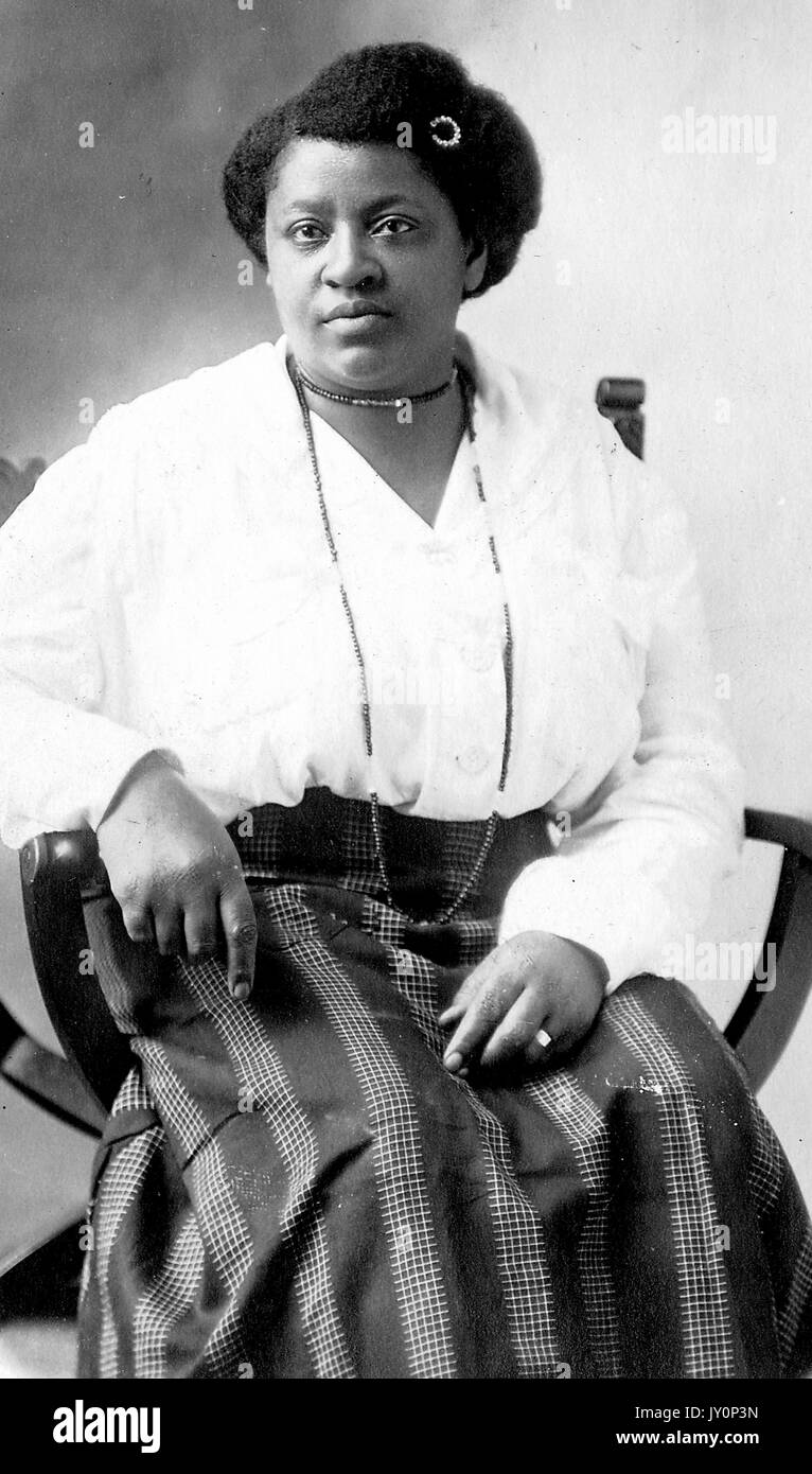 Three quarter length seated portrait, mature African American women, wearing dark skirt with patterns, light blouse, necklaces and hairpin, neutral expression, 1920. Stock Photo