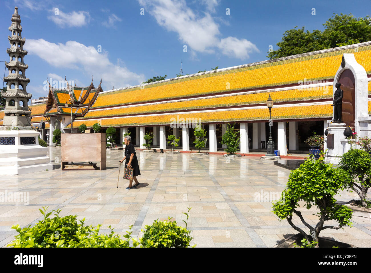 Old woman with walking stick walking across the courtyard at Wat Suthat Thepwararam, Bangkok, Thailand Stock Photo