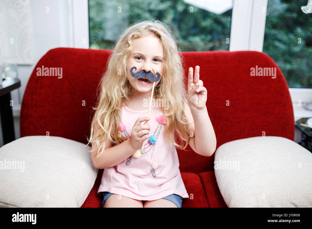 Cute little girl with paper mustaches while sitting on red chair at home. Stock Photo