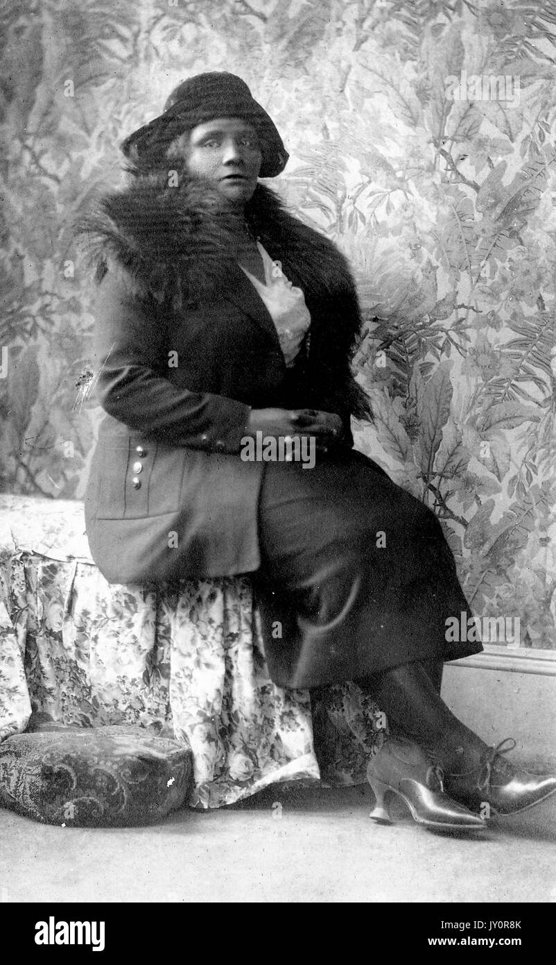 Full length sitting portrait, a mature African American woman, wearing dark dress, overcoat, fur shawl, and hat, floral wallpaper in background, serious expression, 1920. Stock Photo
