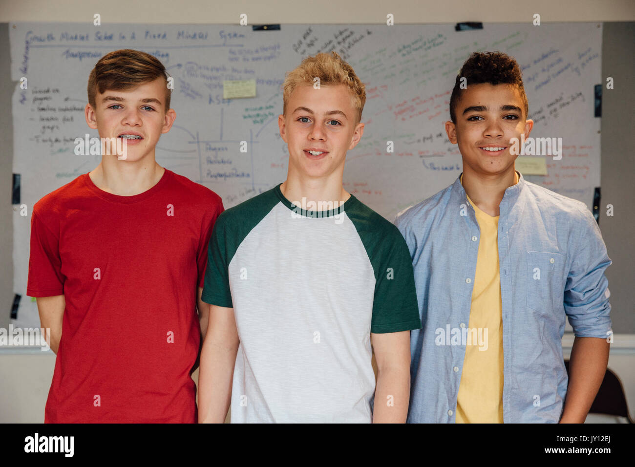 Three male teenagers are posing for the camera at school. They are standing in front of the whiteboard and are smiling at the camera. Stock Photo