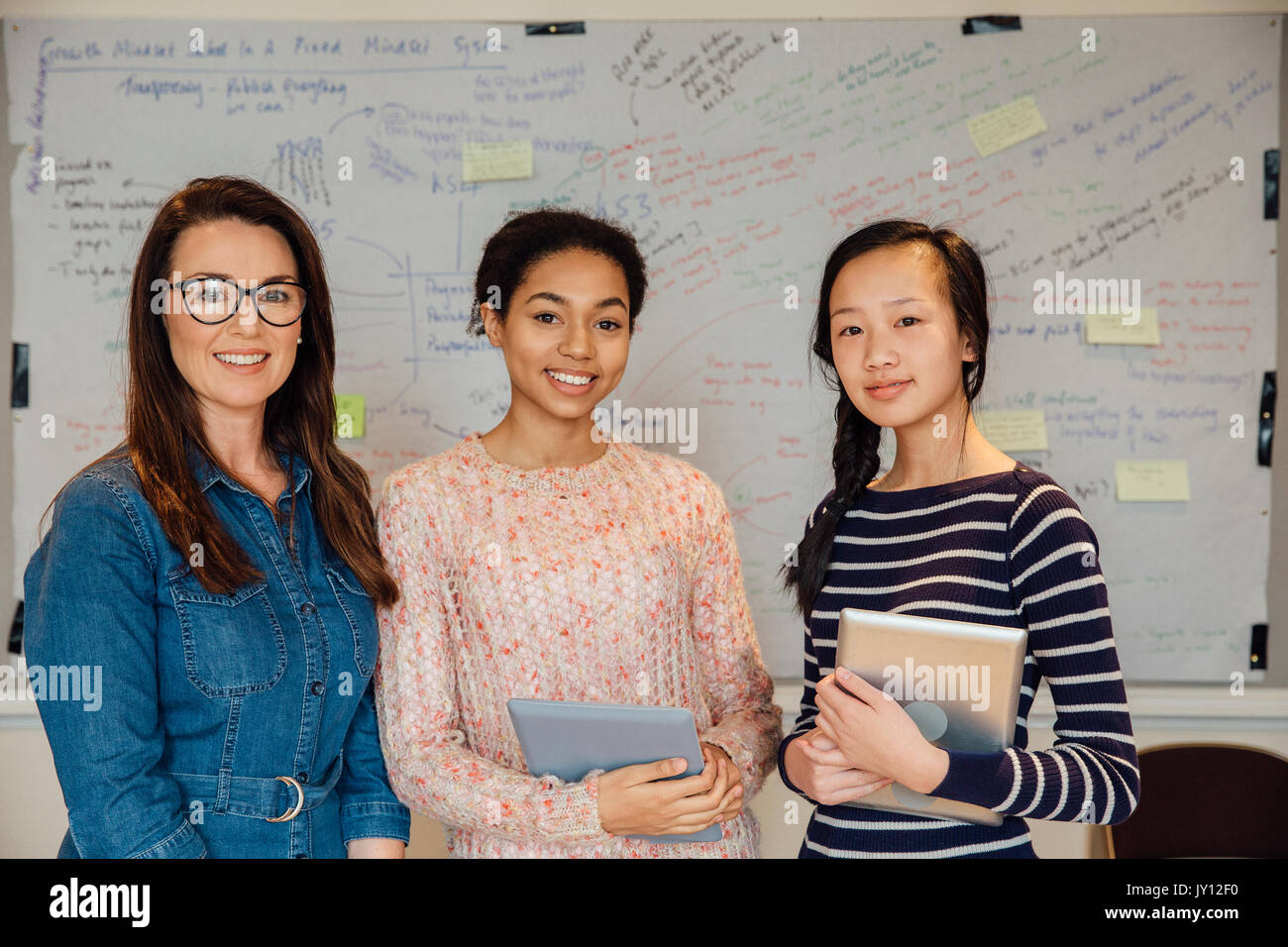 Female teacher and two of her teen students are smiling for the camera in front of the whiteboard in a school classroom. The students are holding digi Stock Photo