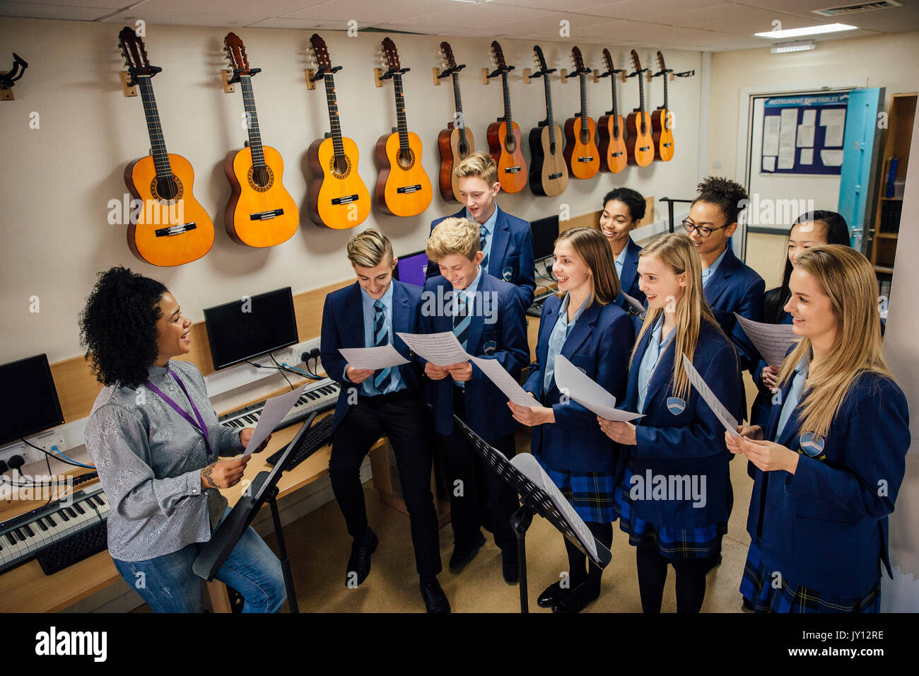 Class of teens are in their music lesson at school. There is a female teacher and the class are practicing choir with her. Stock Photo