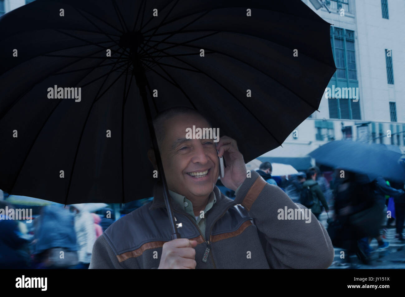 Smiling Hispanic man holding umbrella and talking on cell phone Stock Photo