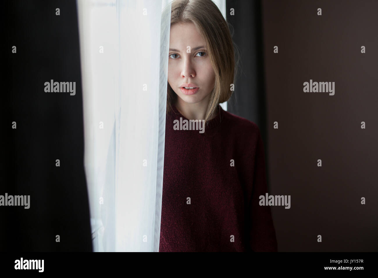 Serious Caucasian woman standing near curtain at window Stock Photo
