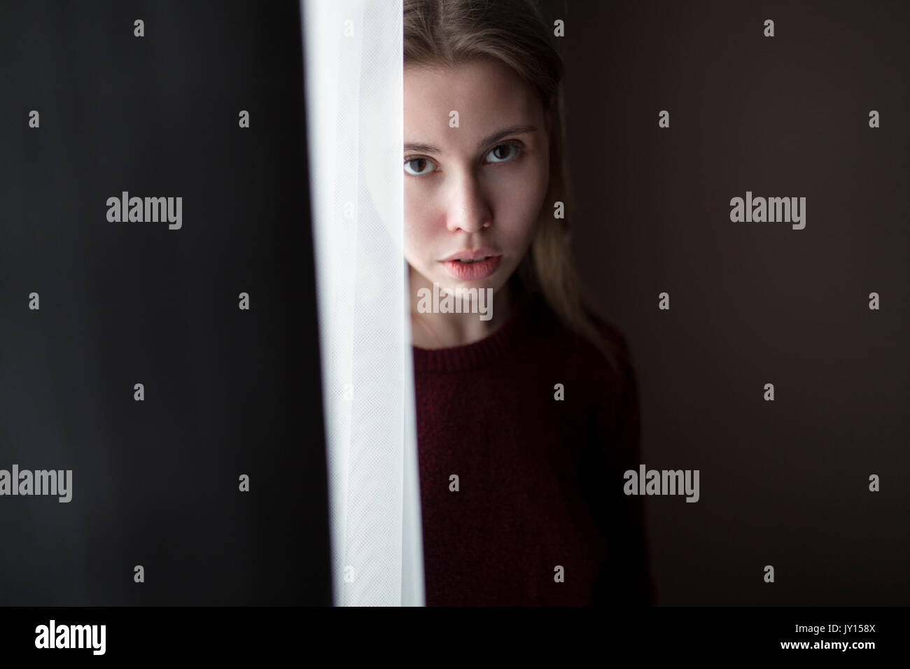 Serious Caucasian woman standing near curtain at window Stock Photo