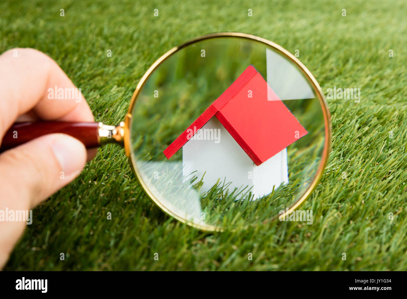 Person Hands With Magnifying Glass Inspecting A Model House On Grassy Field Stock Photo
