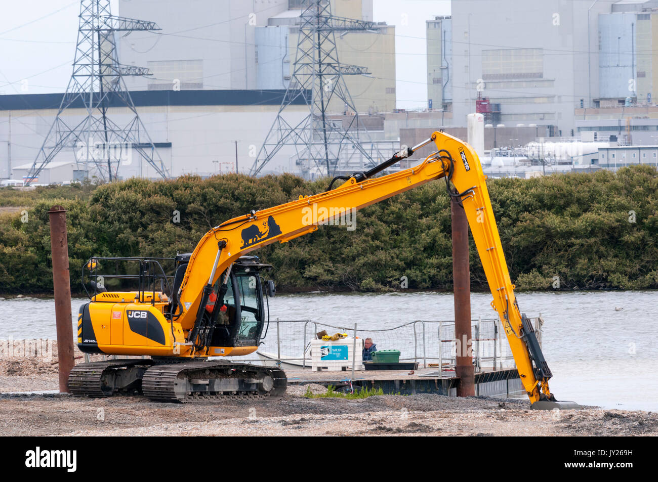 JCB JS145 Tracked Excavator working in a flooded gravel pit at RSPB Dungeness, in front of Dungeness nuclear power station. Stock Photo