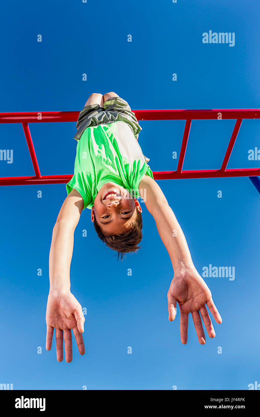 A happy young boy hangs upside down from the monkey bars Stock Photo