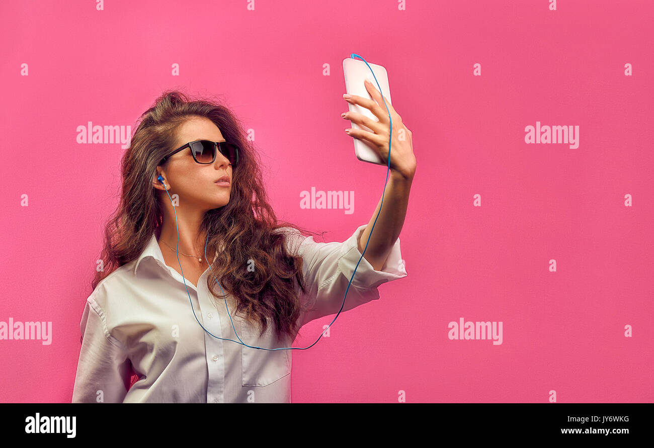 Beautiful caucasian woman in a white shirt  in glasses with headphones, holding a white tablet in hands and makes selfie photo on a pink bright  backg Stock Photo