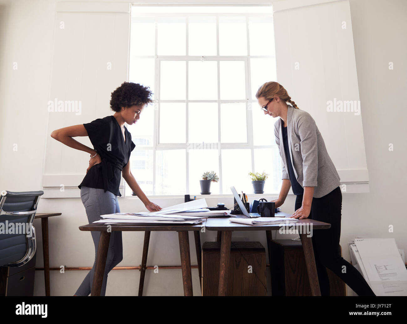 Two women stand working at opposite sides of an office desk Stock Photo