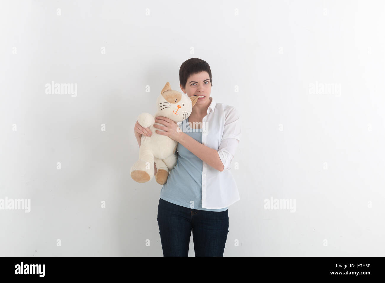 Furious woman bite her plush toy cat for ear, gray studio background. Funny situation. Stock Photo