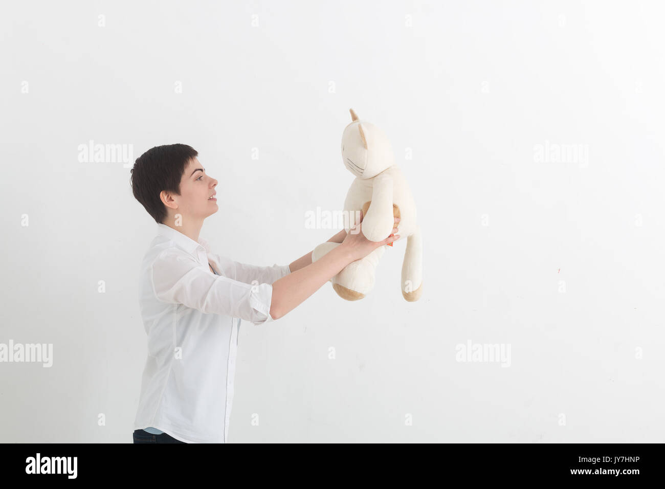 Strange young woman with short hair in white shirt talking to her toy plush cat on light grey background. Stock Photo