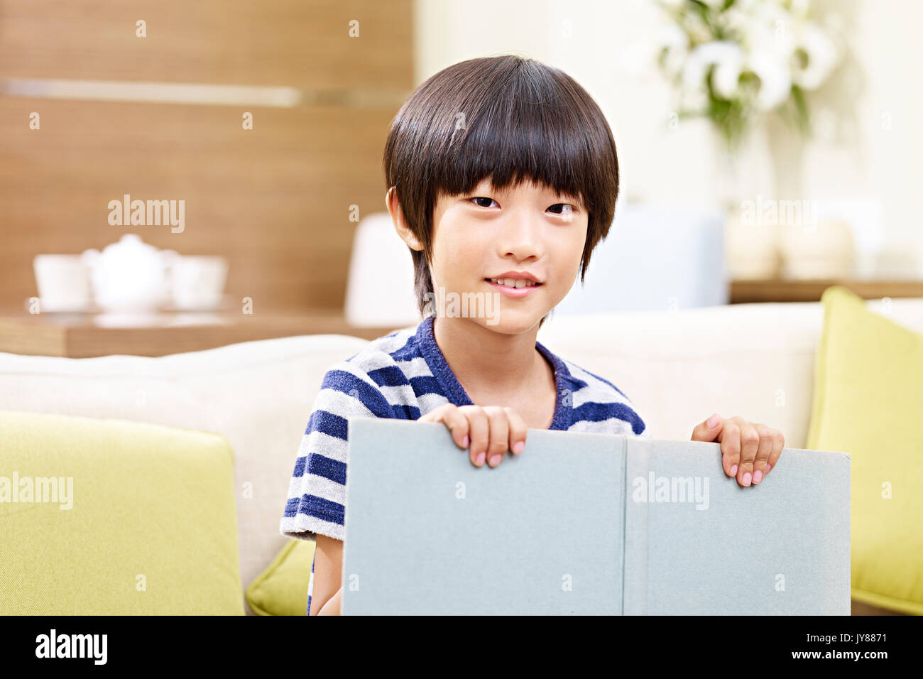 portrait of an asian little boy sitting on couch at home holding a book looking at camera smiling. Stock Photo