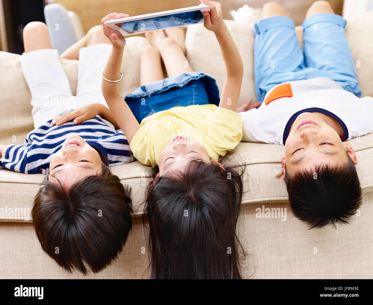 three asian children lying on couch at home playing video game using digital tablet. Stock Photo
