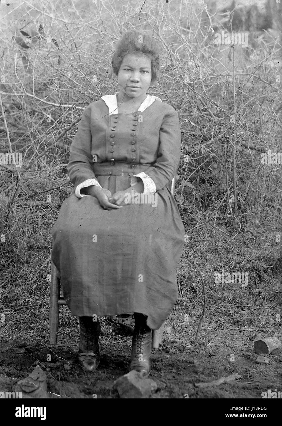 Full length seated portrait of young African American woman, wearing a dress, lace up boots, and a neutral expression, sitting outside in front of plants, 1915. Stock Photo