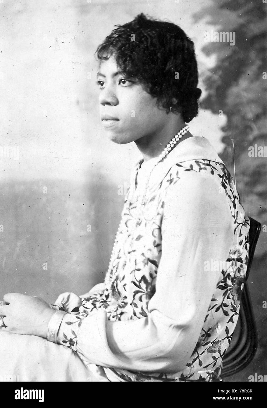 Half length seated portrait of young African American woman, wearing a pattered garment, a necklace, a bracelet, and a neutral expression, 1915. Stock Photo