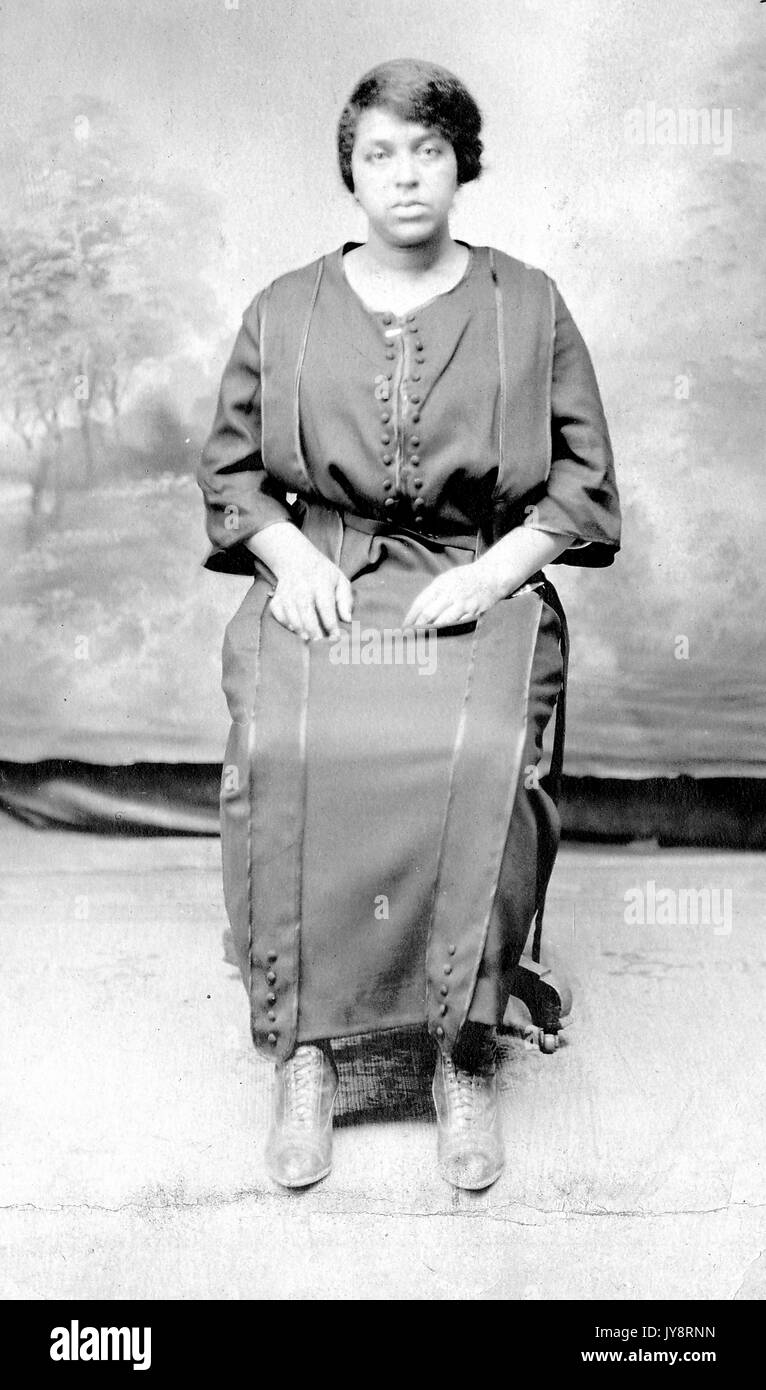 Full length seated portrait of young African American woman in front of a painted background, wearing a dark dress, lace up shoes, and a serious expression, 1915. Stock Photo