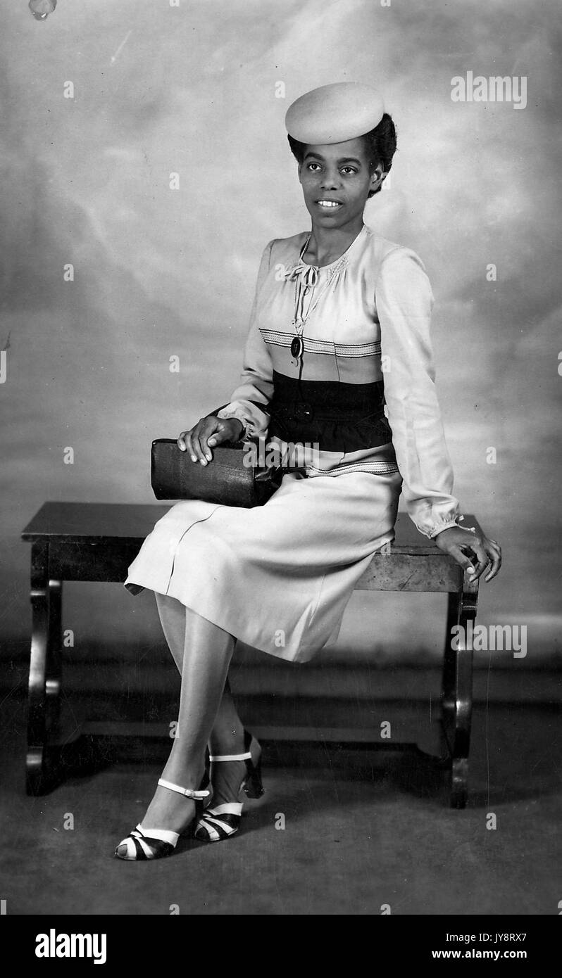 Young African American woman in elegant clothing sitting on a bench and posing for a studio portrait, with a hat and purse, Kentucky, 1920. Stock Photo