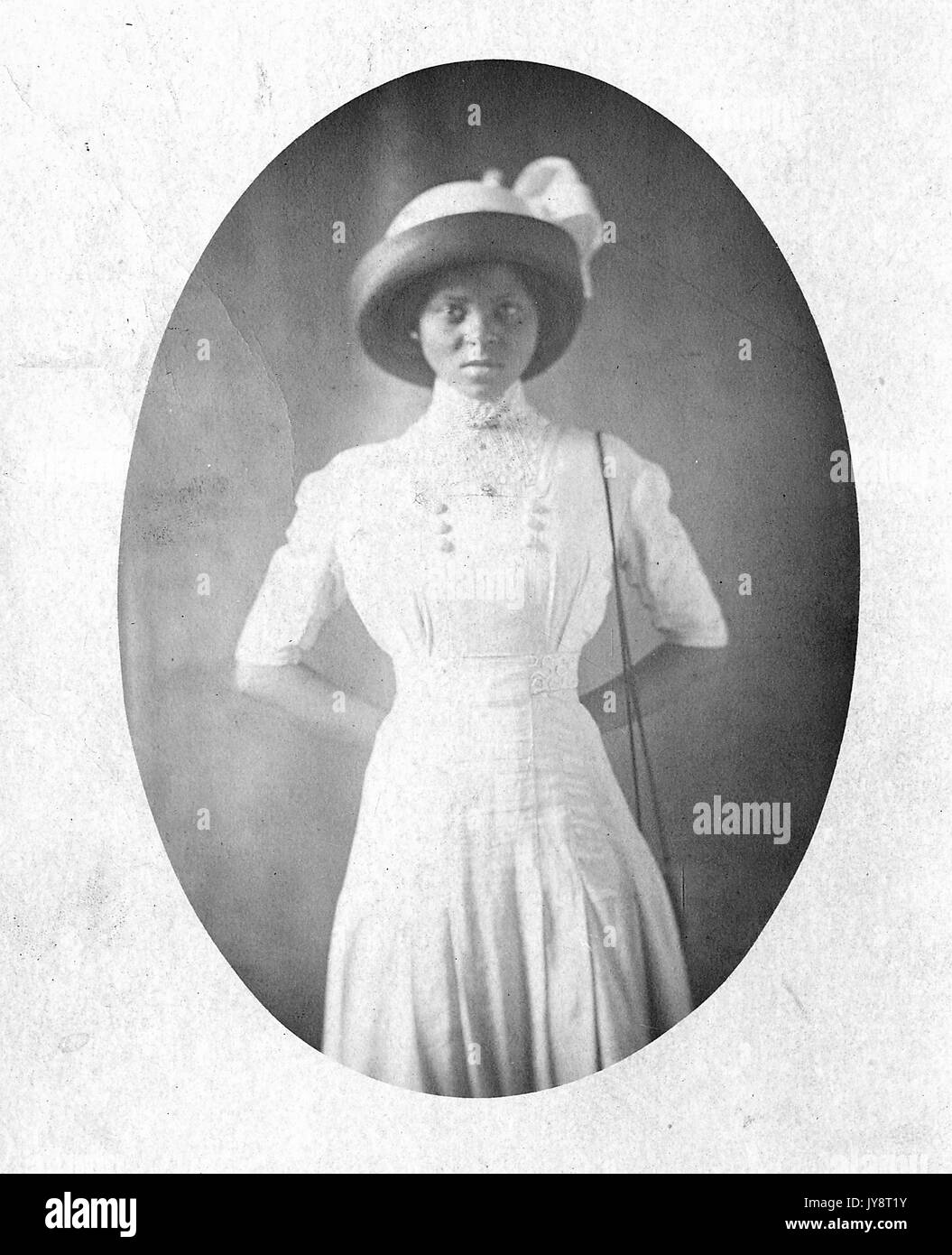 Three quarters length portrait of young African American woman with formal hat and white dress, her hands clasped behind her back, with a serious facial expression, 1915. Stock Photo