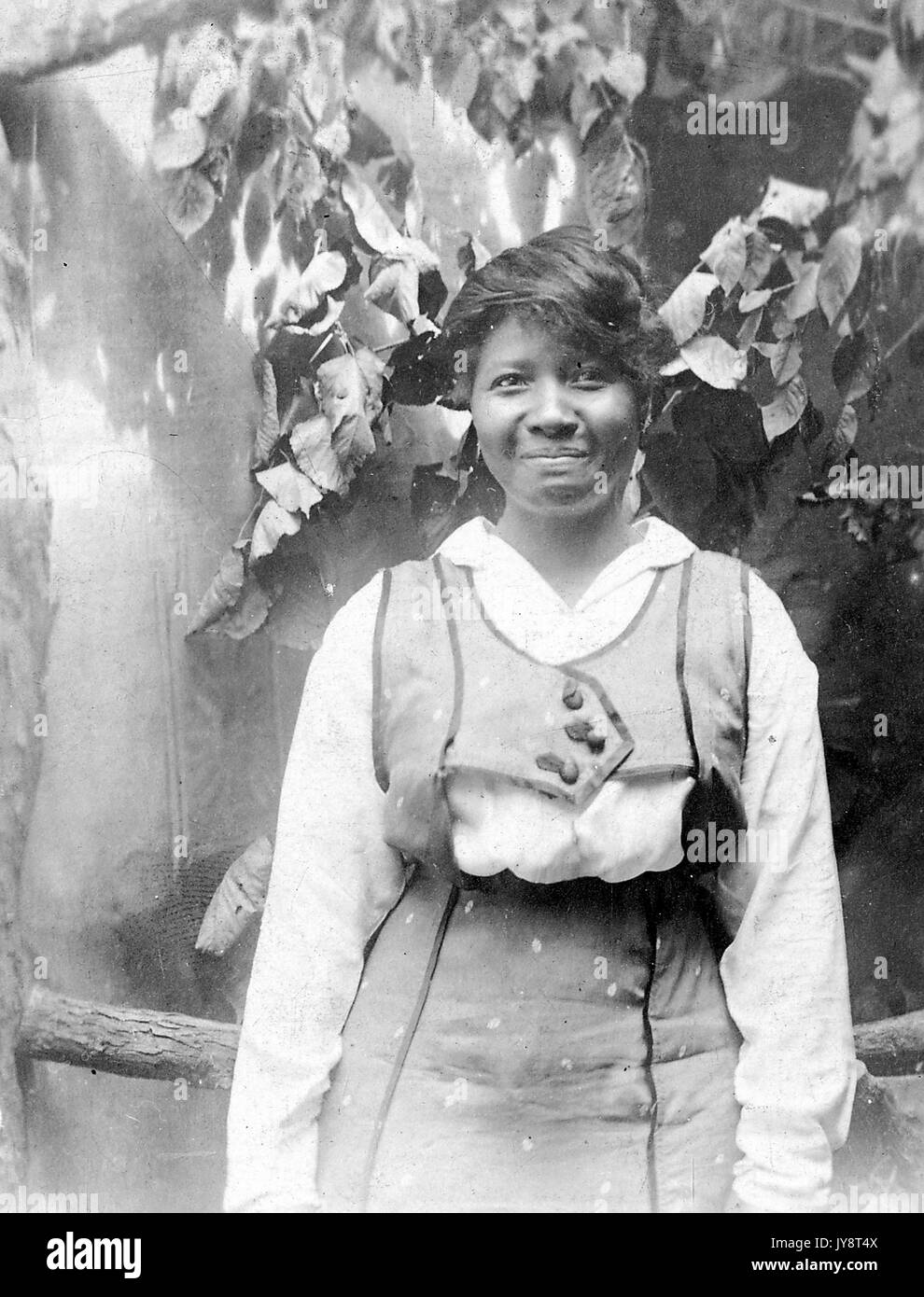 Half length portrait of African American woman standing in front of a painted backdrop featuring a natural scene, with a slight smile, 1915. Stock Photo