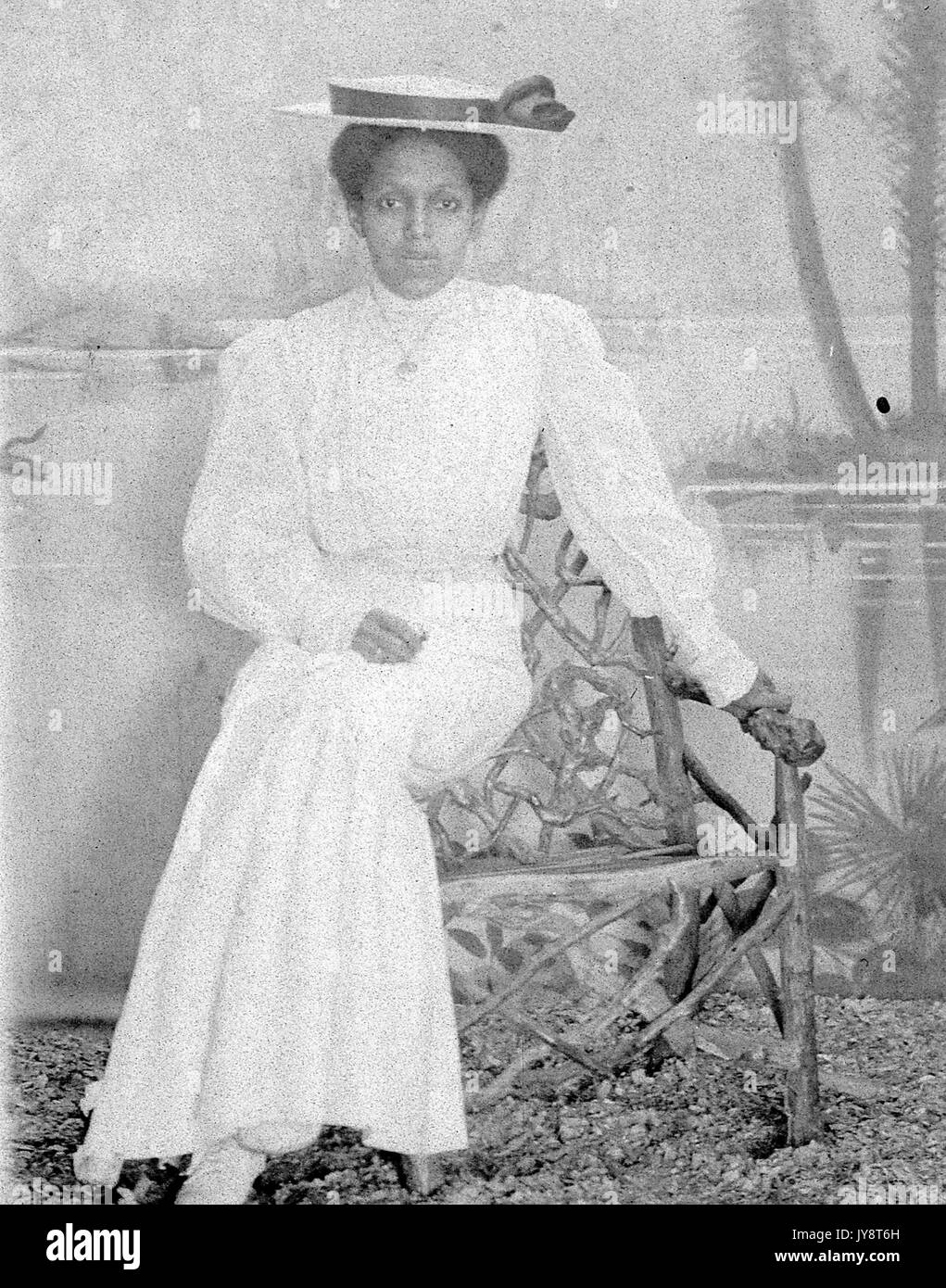 Full length portrait of African American woman wearing a hat and sitting on a handmade chair in a photographic studio, painted backdrop in the background shows a lake scene, 1915. Stock Photo