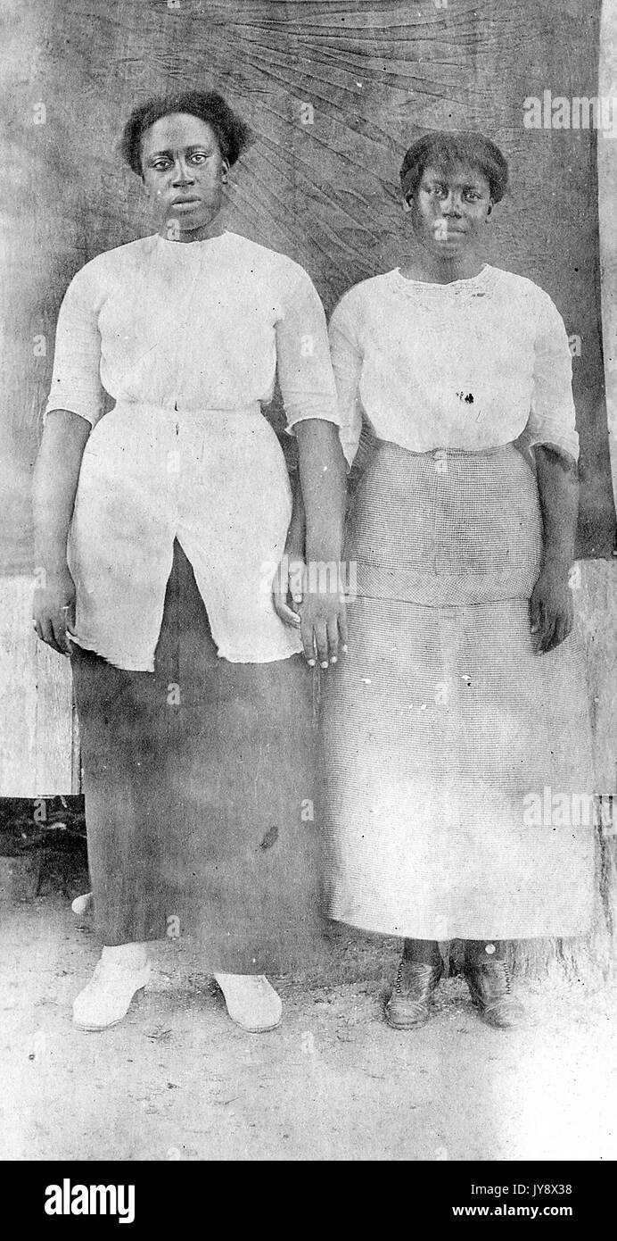 Full length portrait of two African American Women, side by side with arms and hands touching, the one on the left in a dark dress, the other in a light dress, in front of a dark backdrop, both with serious facial expressions, 1920. Stock Photo