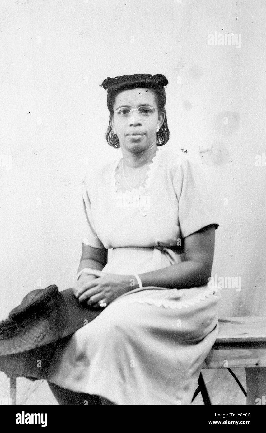 Three quarter length portrait of African American woman, sitting, hands folded in lap, hat and coat in lap, wearing a lace trim dress, neutral facial expression, 1920. Stock Photo