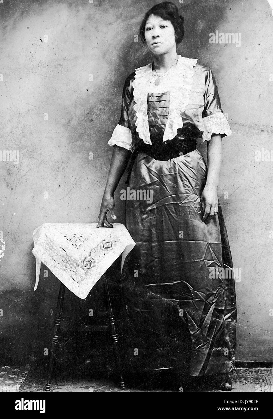 Full length standing portrait of mature African American woman, wearing dark dress with lace design and trims, standing next to stool covered in lace cloth, neutral expression, 1915. Stock Photo