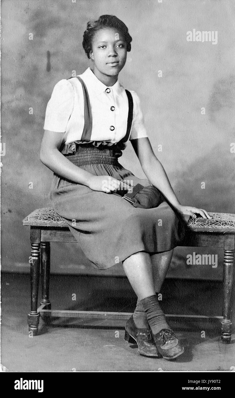 Portrait of an African American woman sitting on a bench, wearing a white button down shirt and a high-waisted skirt, holding a purse, 1915. Stock Photo
