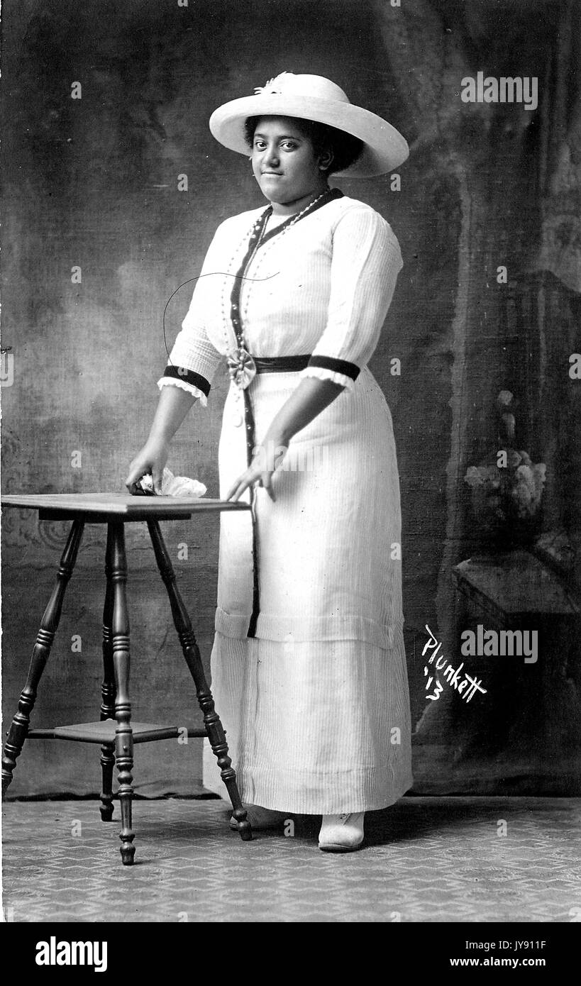 African-American woman, full length portrait, wearing white dress and large hat, standing behind wooden stool, with a slight smile, June, 1913. Stock Photo