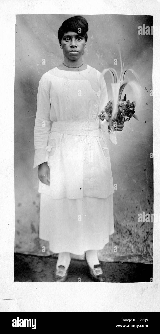 Full length standing portrait of mature African American woman with neutral expression, wearing light dress, necklace, and buckled shoes, holding plants, 1915. Stock Photo
