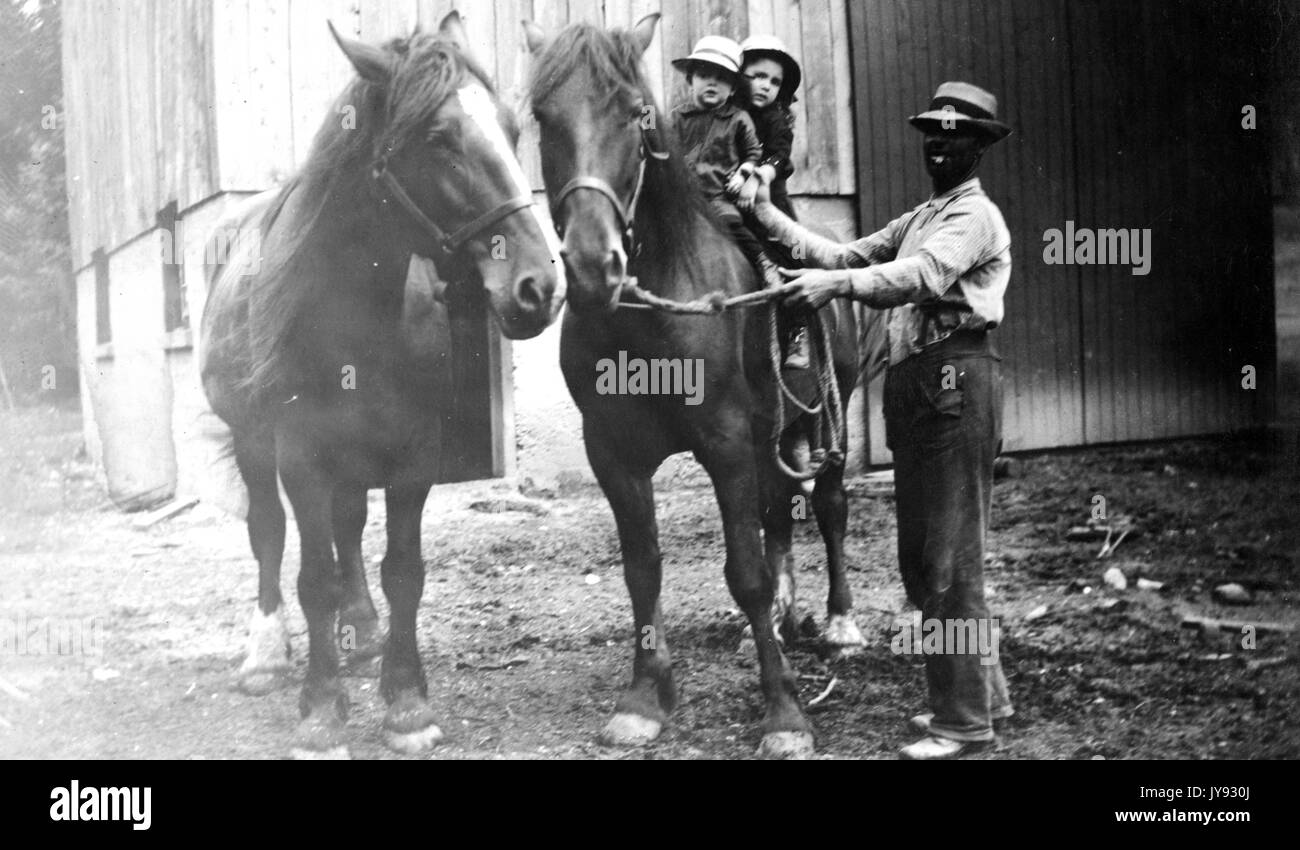 African-American man helping two young Caucasian children onto the back of a large draft horse, the man smiling and holding the reins of the horse, 1930. Stock Photo